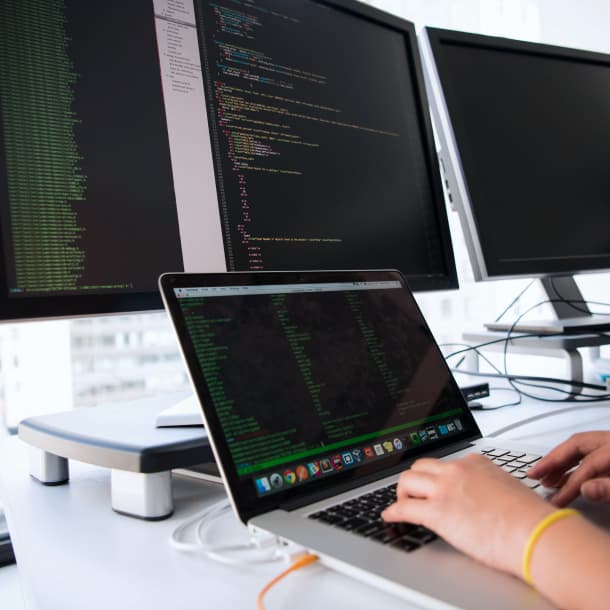 A person writing code on a laptop next to three monitors