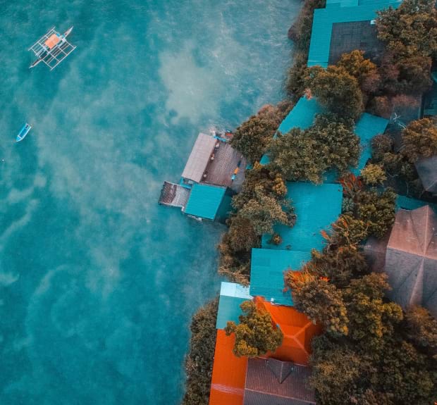 Aerial view of a house situated on the water, surrounded by greenery and reflecting the sky in the calm surface below.